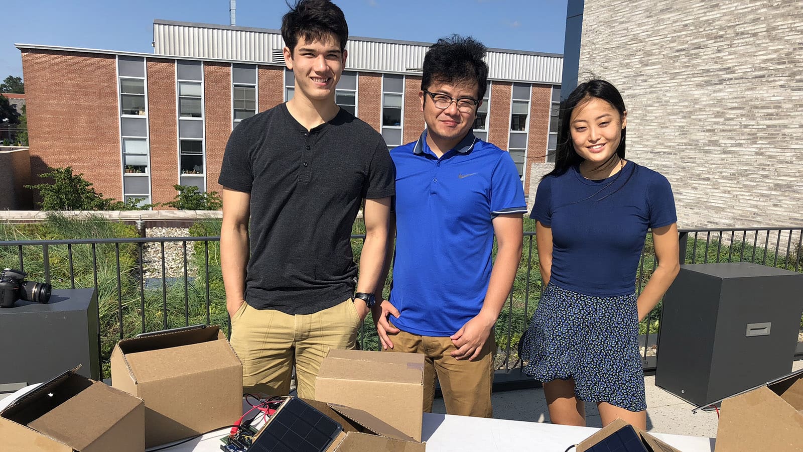 Three students stand on rooftop with small solar panels