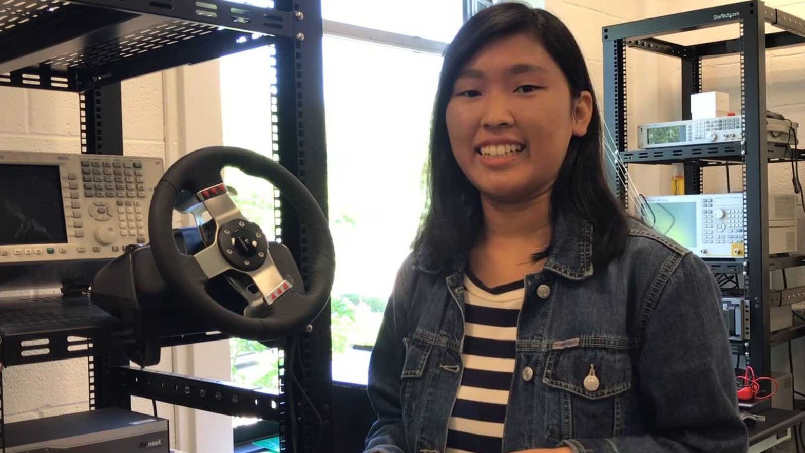 Surin Ahn smiles in the lab, with a steering wheel attached to a computer in the background.