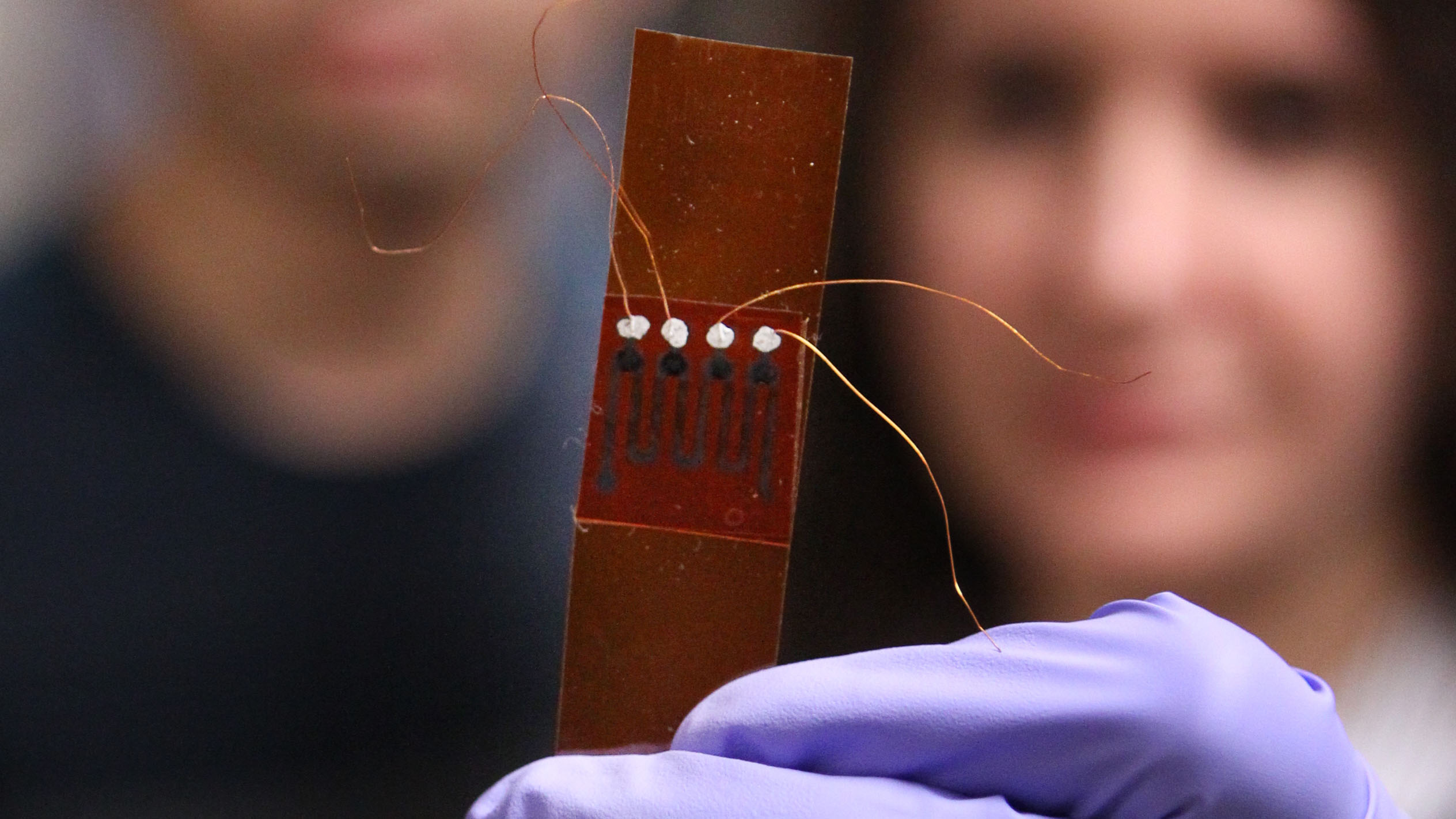Researchers, out of focus, hold photovoltaic device close to the camera.