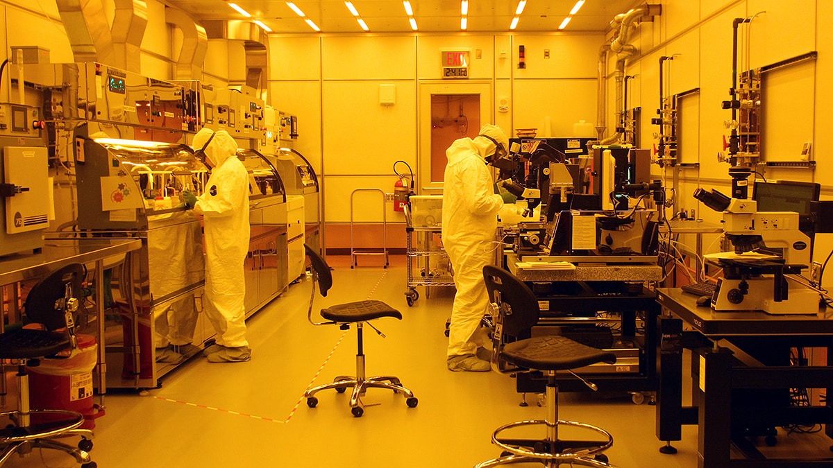 Researchers, dressed in white suits, work in "clean room" lab under yellow light.