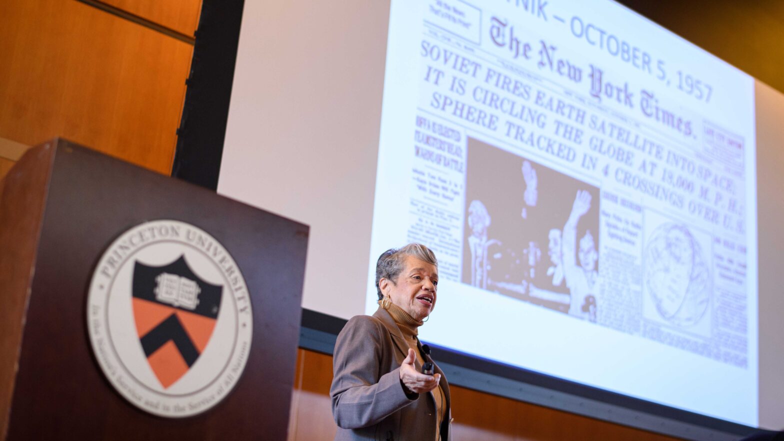 Woman speaking next to a lectern with projected slide showing front page of The New York Times