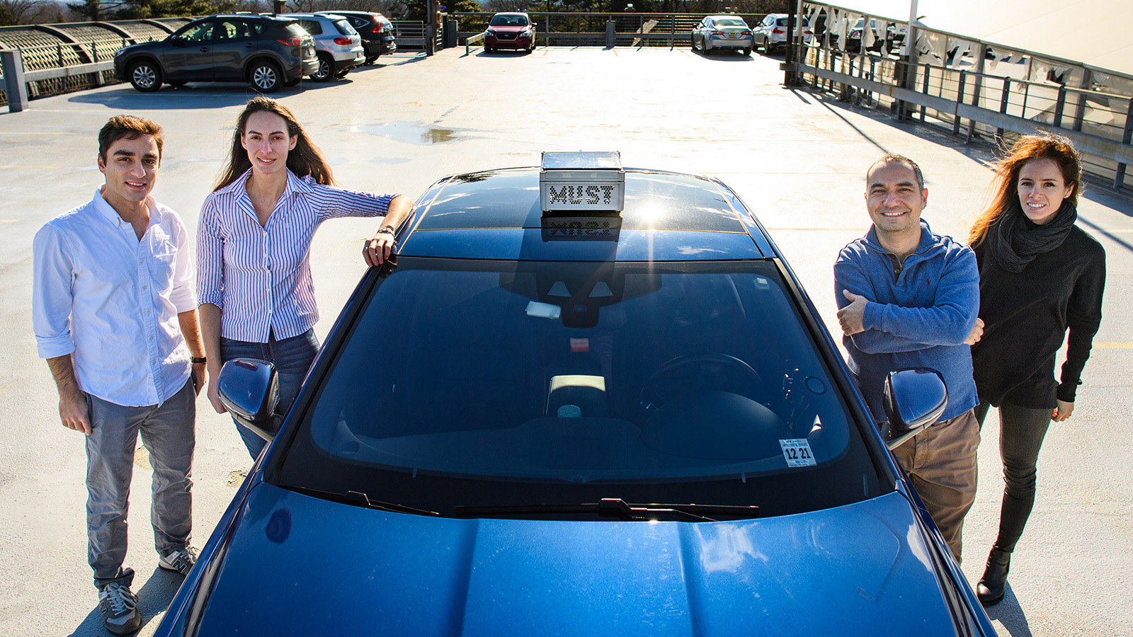 Four researchers stand next to a blue car, two people on each side of the car, which has a device on the roof labeled "MUST"