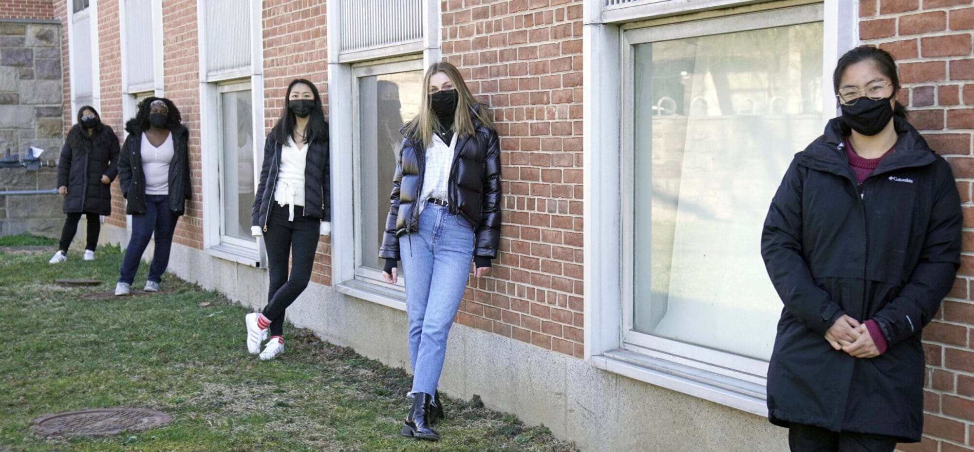Five students in masks leaning against Engineering building, six feet apart.