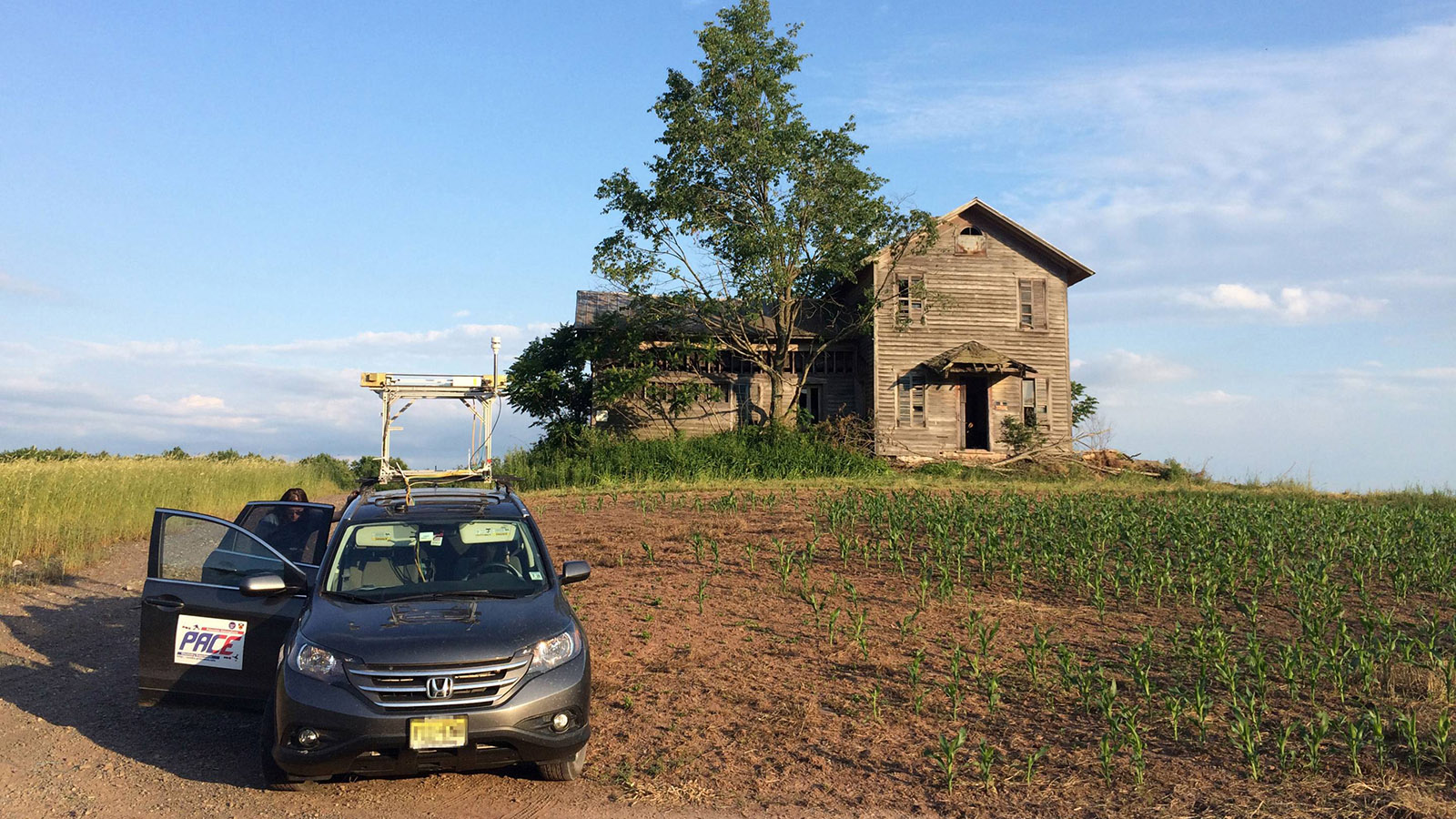 Car mounted with sensing equipment sits in field in front of weathered farmhouse