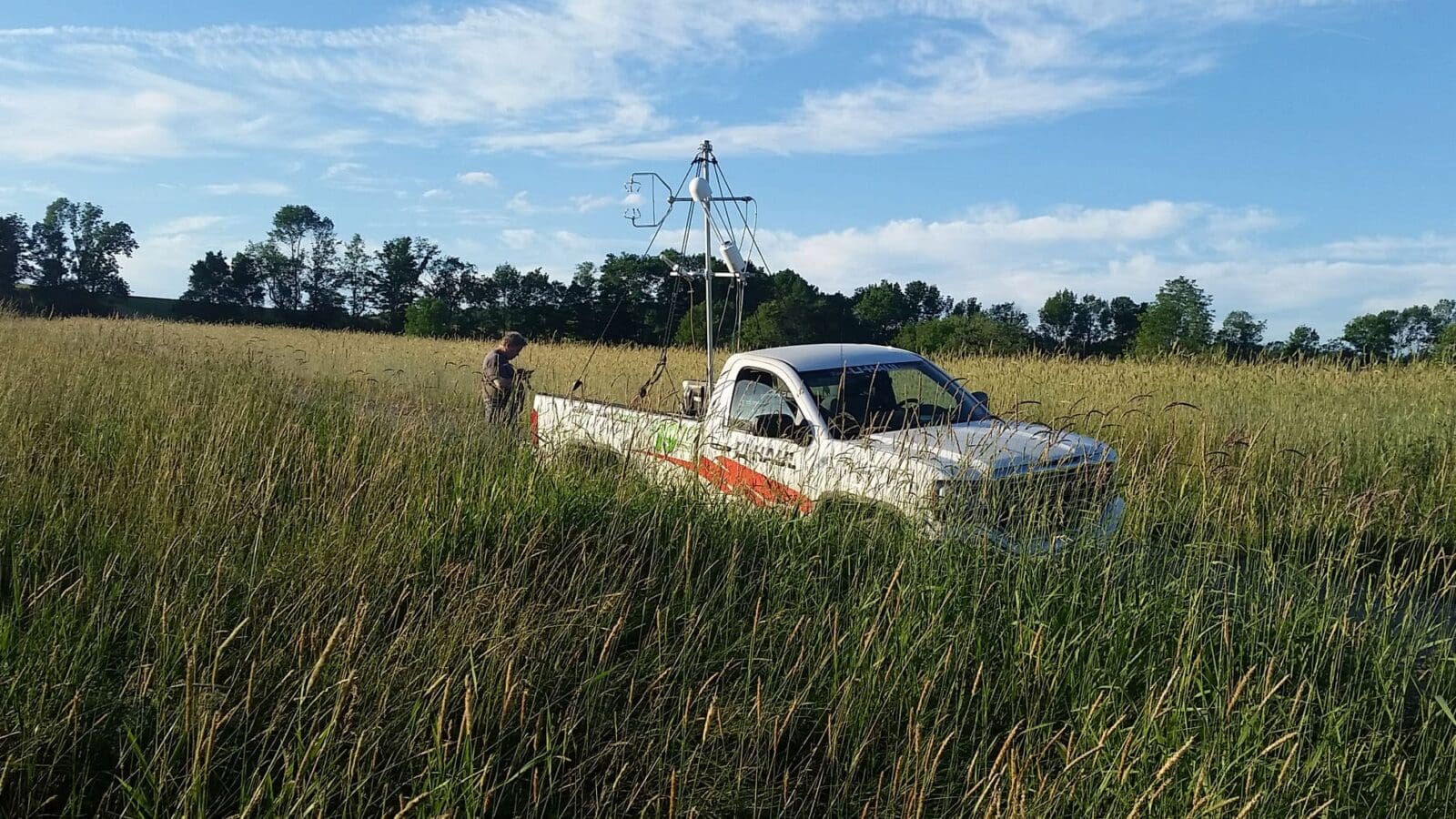 Pickup truck parked in middle of field of tall grass, tall piece of equipment in the bed of track, researcher standing behind the truck. Row of trees and open sky in the background.