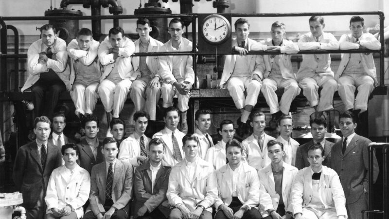 Older black and white photo of young men posing for class picture. The clock says 12:10 p.m.