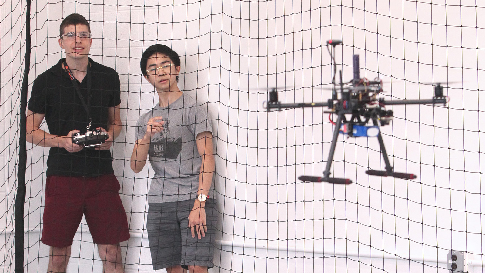 Two students behind mesh curtain flow drone, which is in foreground