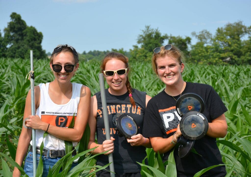Three female students in T-shirts pose with light farming equipment in their hands, smile for camera. They are in a crop field.