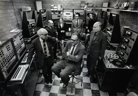 A group of researchers, 1970. perhaps seven, stand in a computer lab. They are dressed in suits. The floor is checkerboard patterned. It is a black and white photo from the 