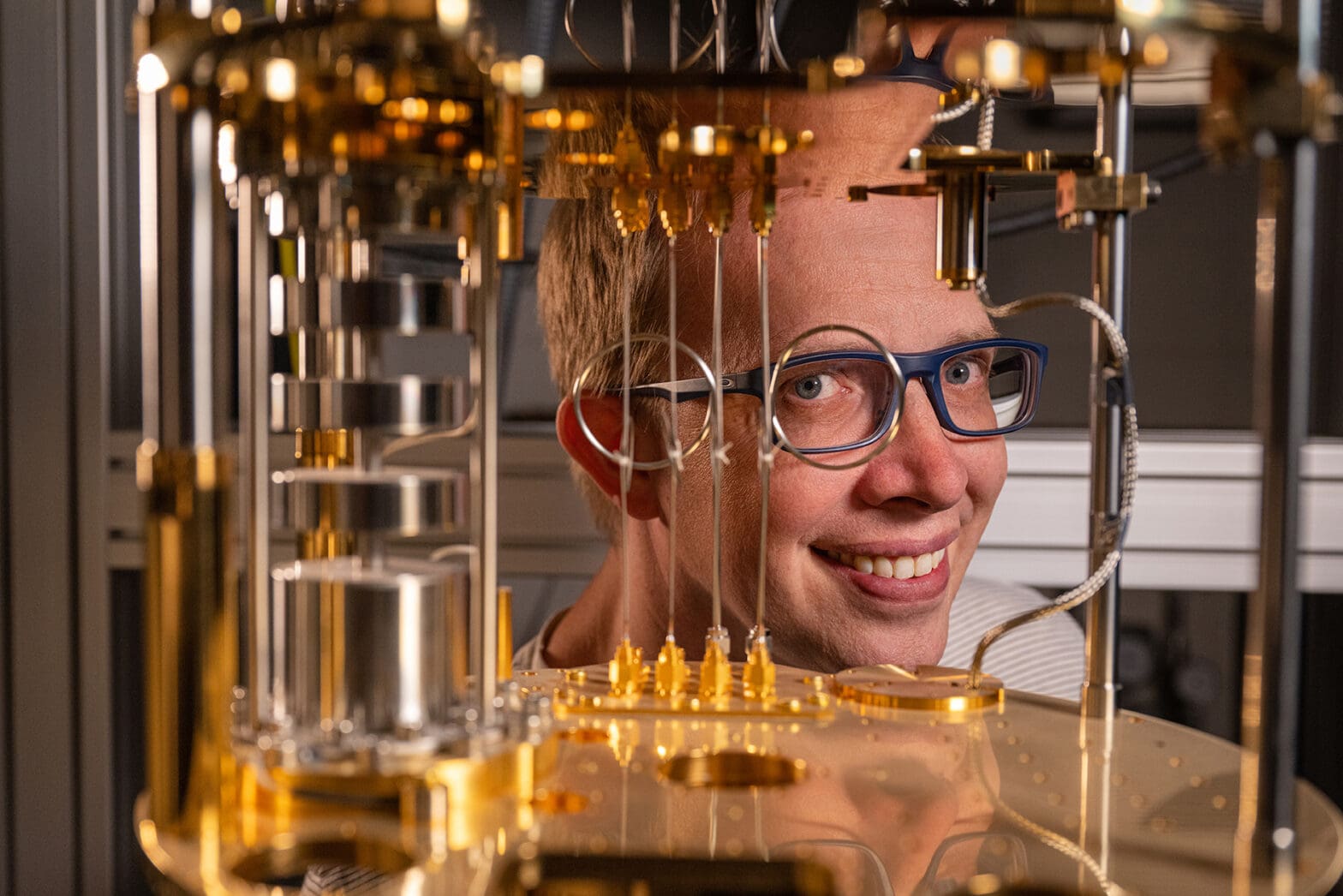 Bespectacled professor smiles and peers through coiled wire between gold discs inside a quantum computer