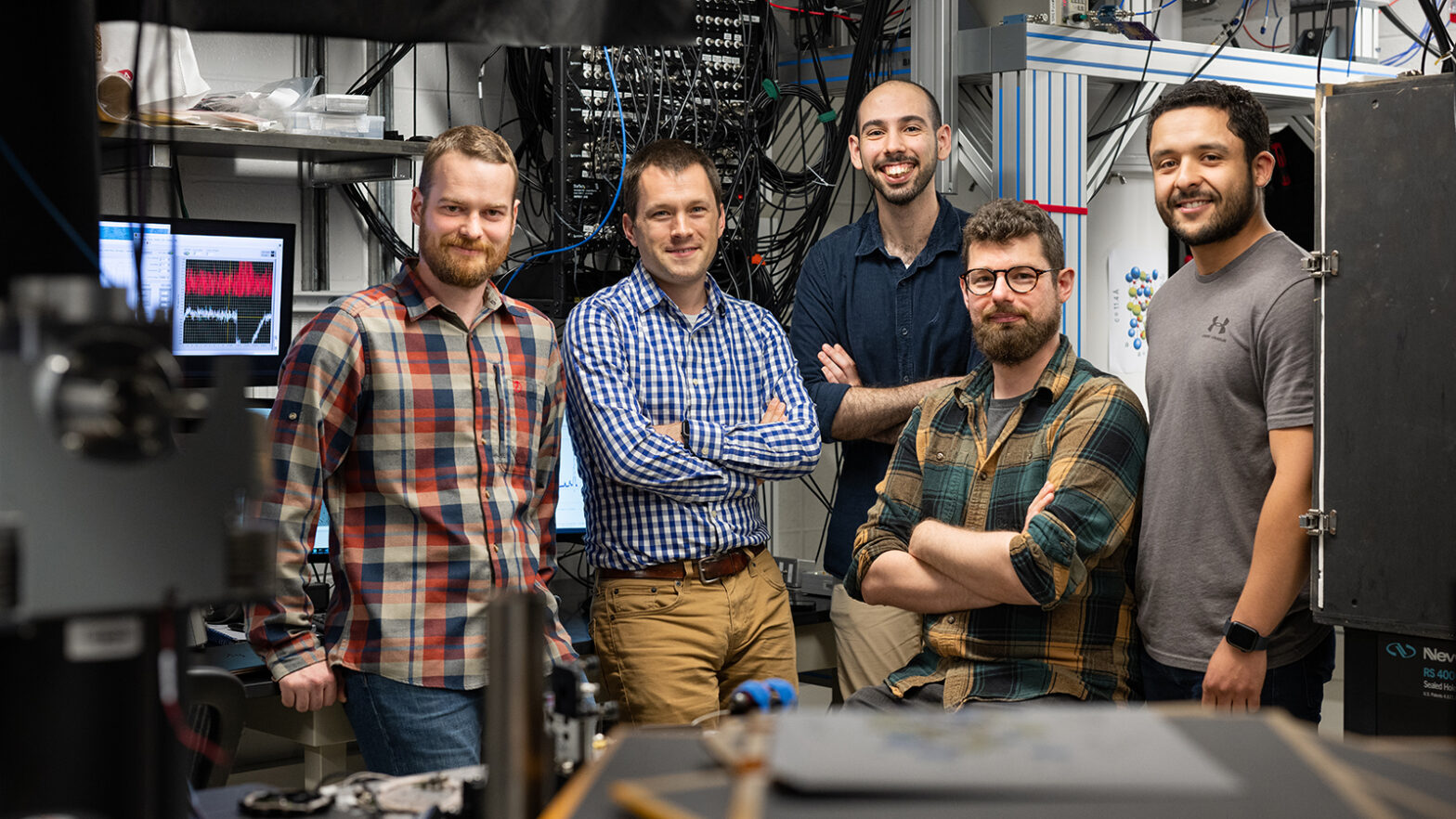 Four graduate students gather around professor Jeff Thompson in the lab, with a quantum computer behind them and figures displayed on the screen.