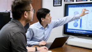 A researcher points at a computer screen while another observes