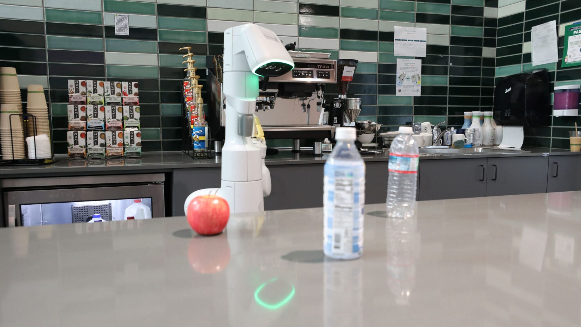 Photo of a robot with a camera "face" looking at a kitchen counter with two plastic water bottles and an apple.