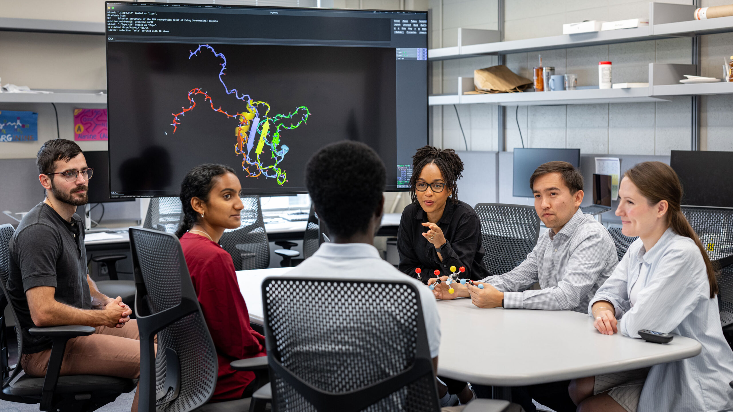 A group of six researchers sits around a table and discusses molecular models, which are visible on a screen behind them and in physical form on the table.