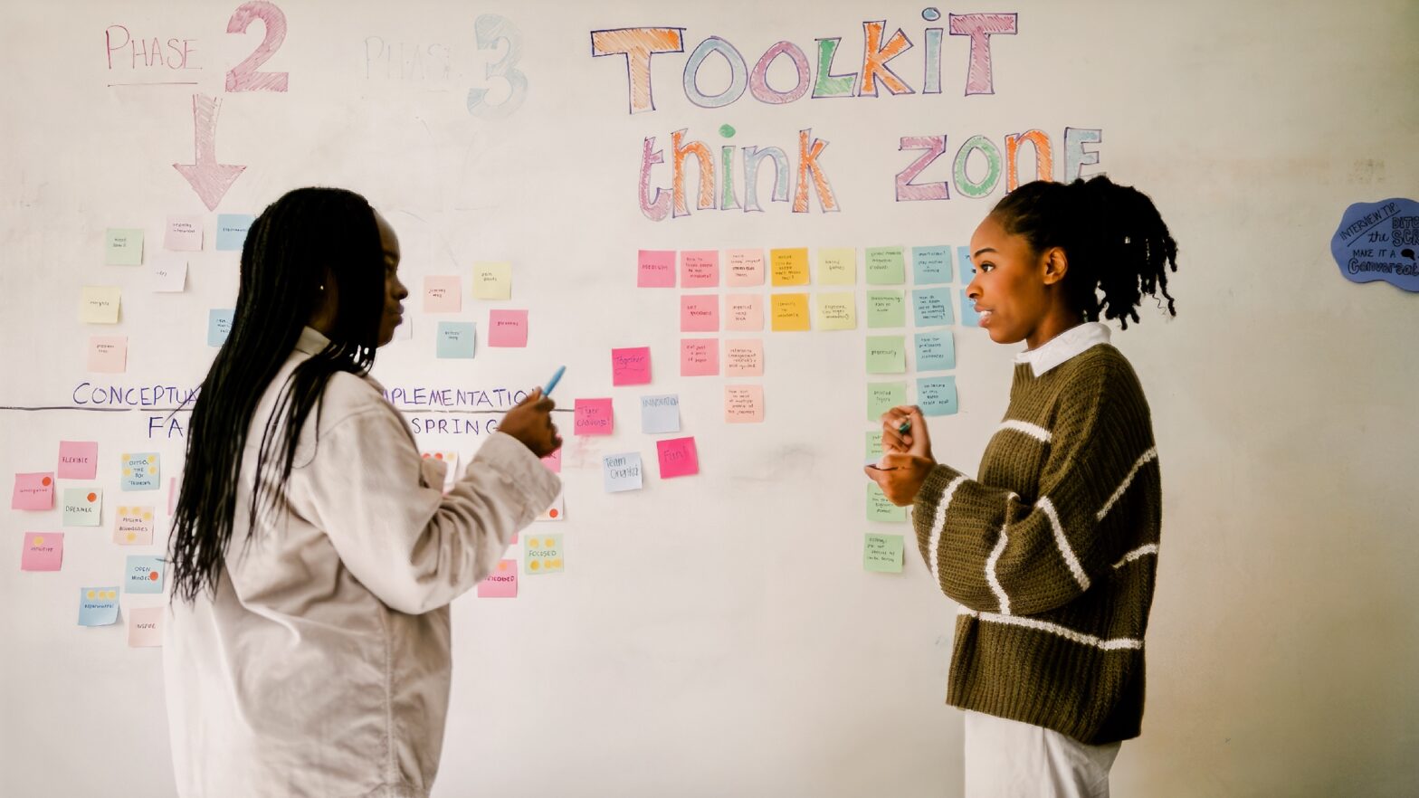 Two students talk in front of large white board covered with sticky notes and writing.