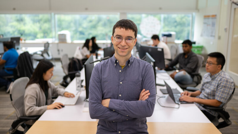 Smiling man with folded arms stands in front of researchers working at computers