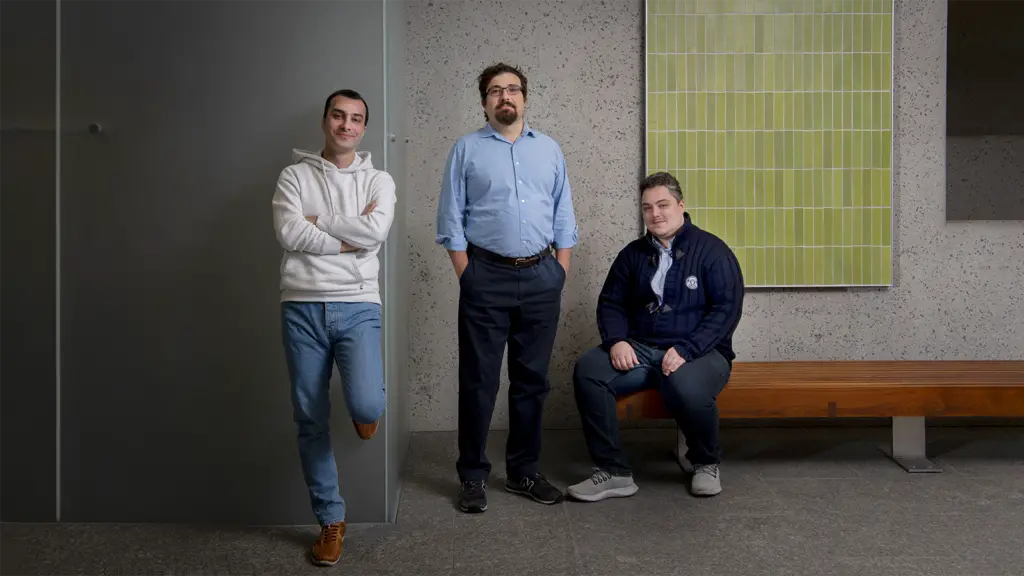 Three men pose in front of a cement wall.