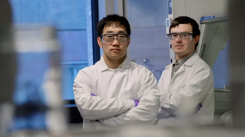 Two men in lab coats and goggles pose in a lab setting.