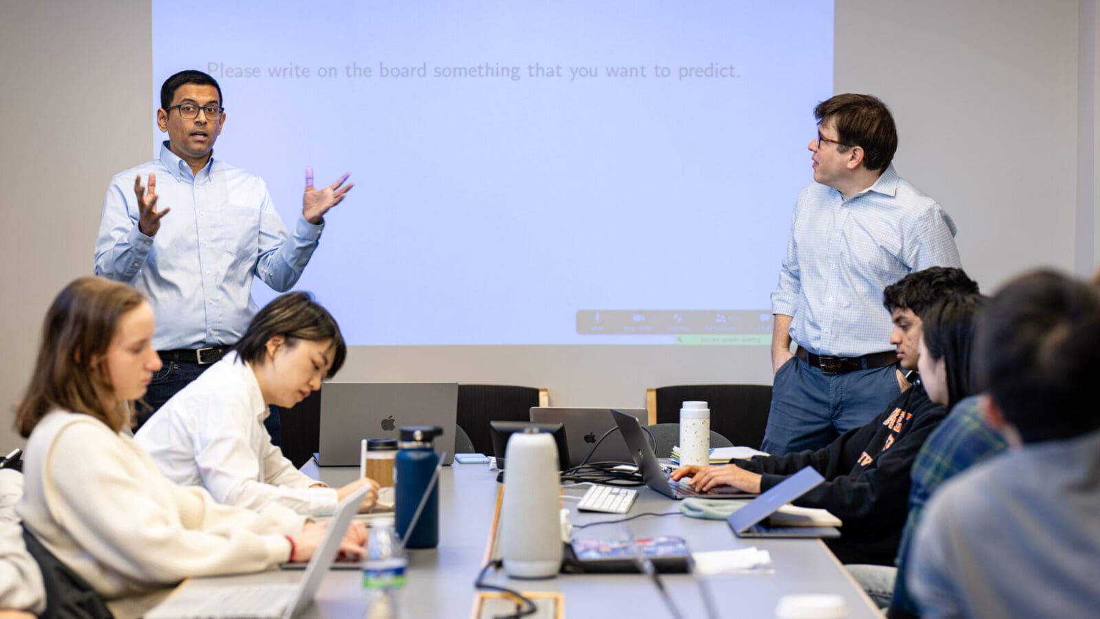 Two instructors speak to students while standing before a white board