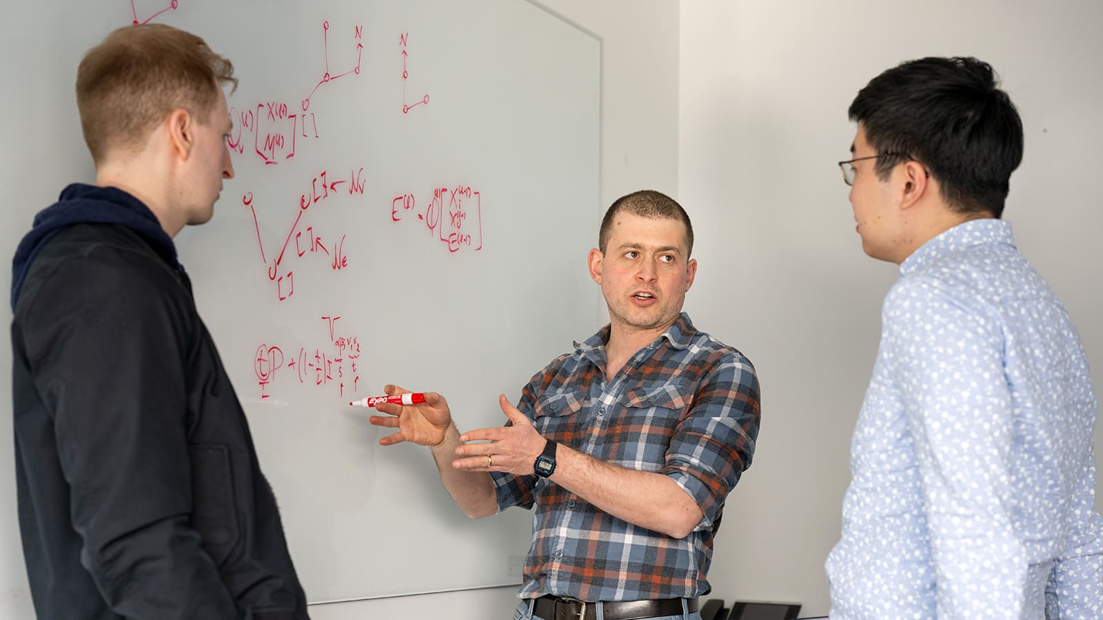 Three researchers stand in front of a white board discussing mathematics written on the board.