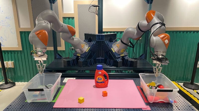 Two robotic arms poised over plastic storage containers of sorted objects. In between the storage containers are additional items to be sorted, including a detergent bottle and two wooden blocks, colored yellow and orange.