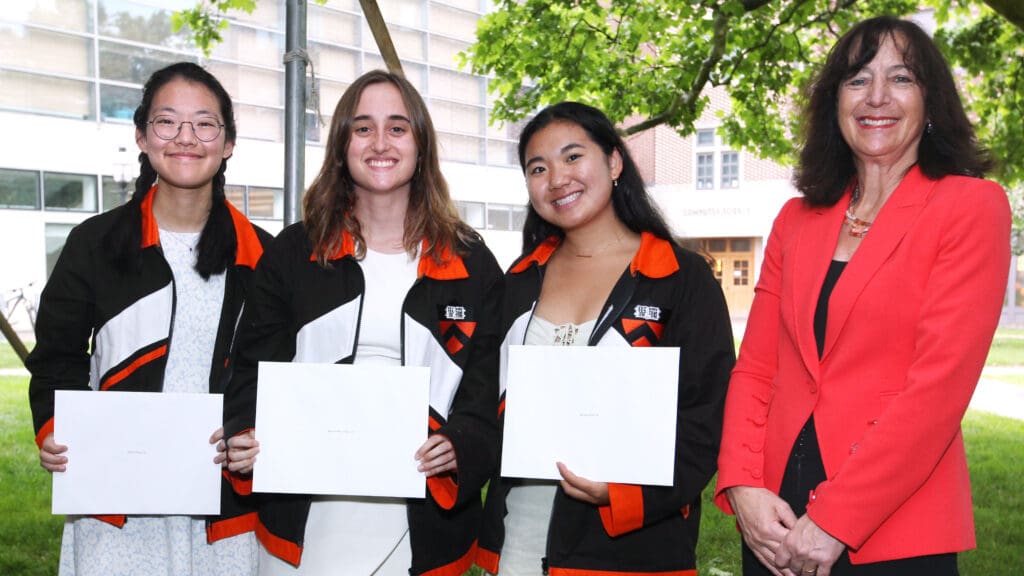 Three women wearing class jackets and holding award plaques stand next to a woman in an orange blazer.