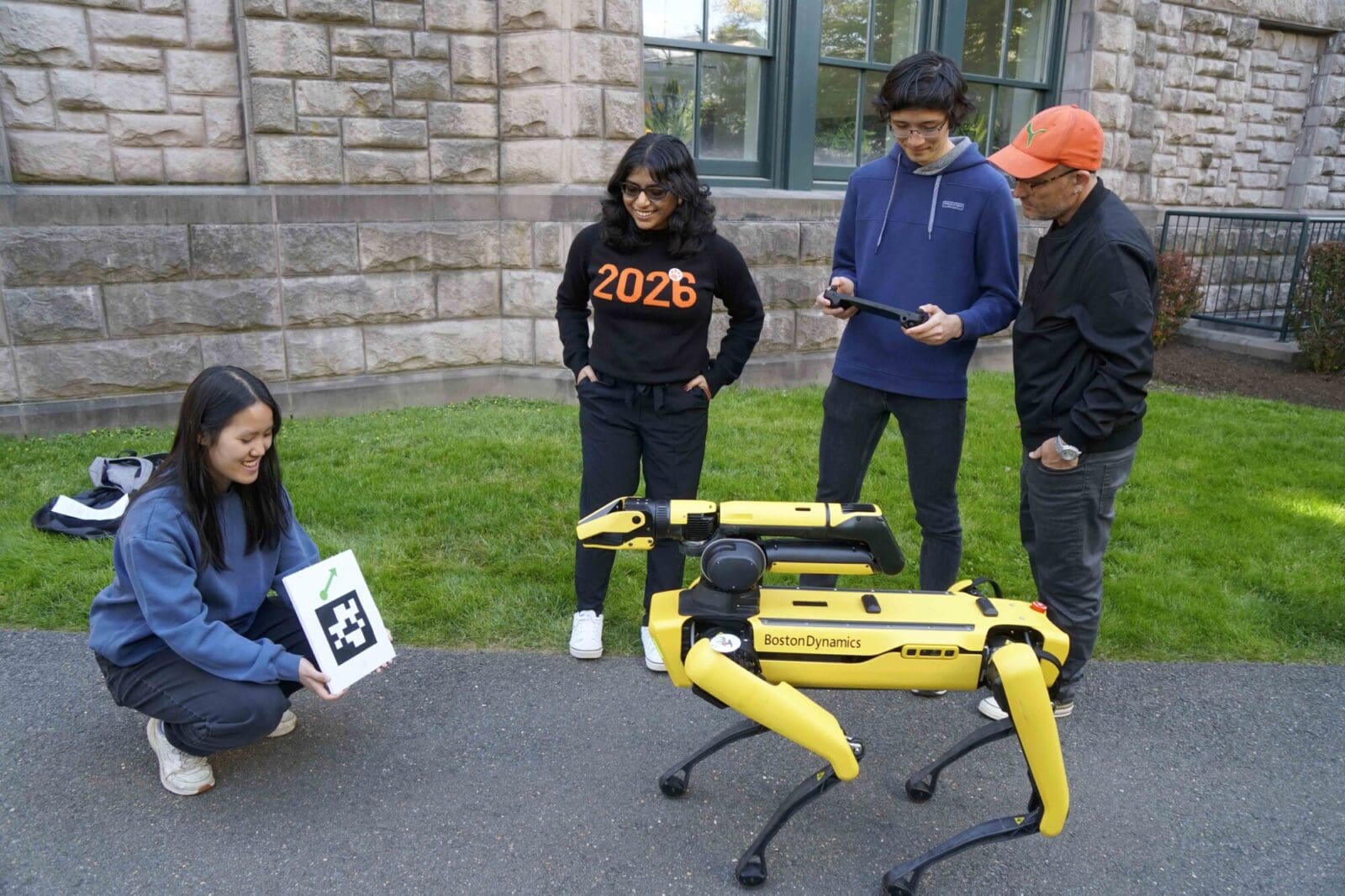 Vivian Chen holds large QR code as Vasumathi Venkat, Aaron Serianni, and Alex Glaser look on, and the robot dog faces the QR code.