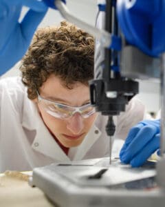 Christopher Browne works a small drill press in lab attire.