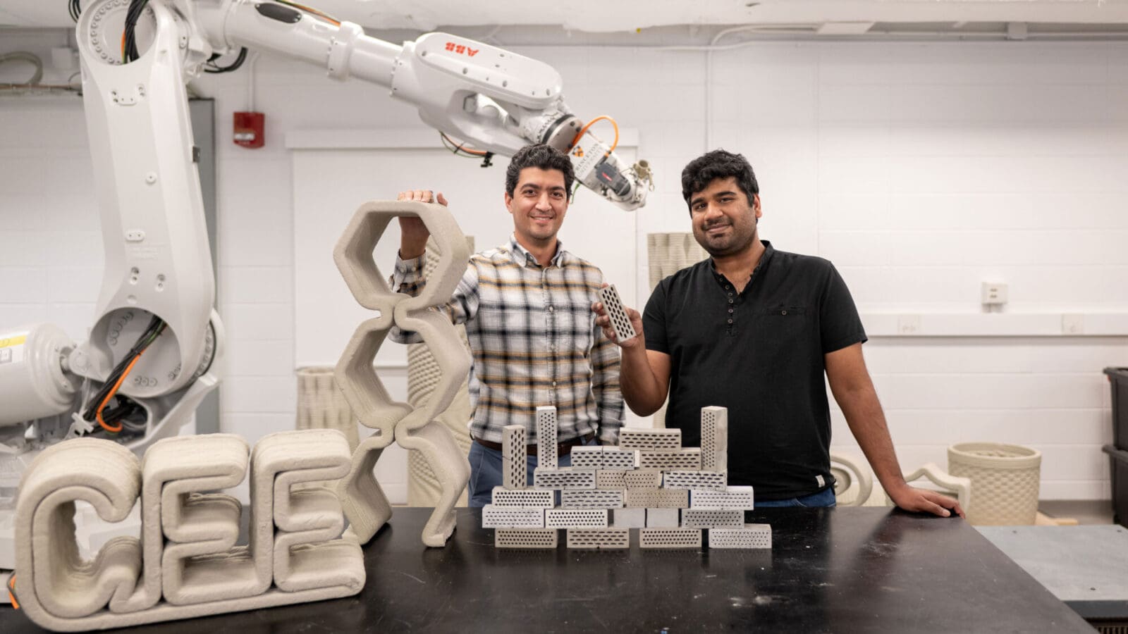 Two men in a lab with an industrial robot and small cement bricks