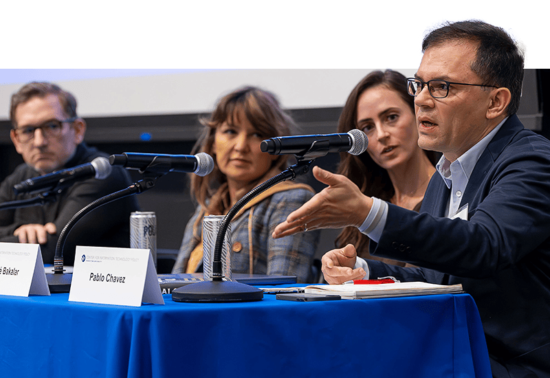 A group of panelists sit behind a table as one man addresses the audience