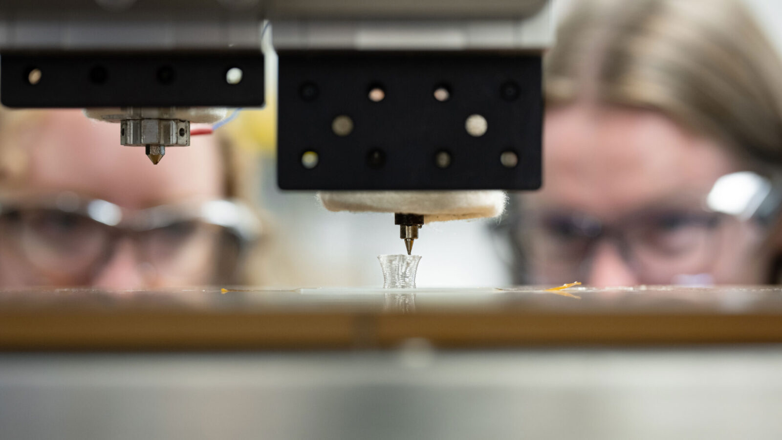 A 3D printer creates a small translucent plastic cylinder. Two blurred faces in the background observe