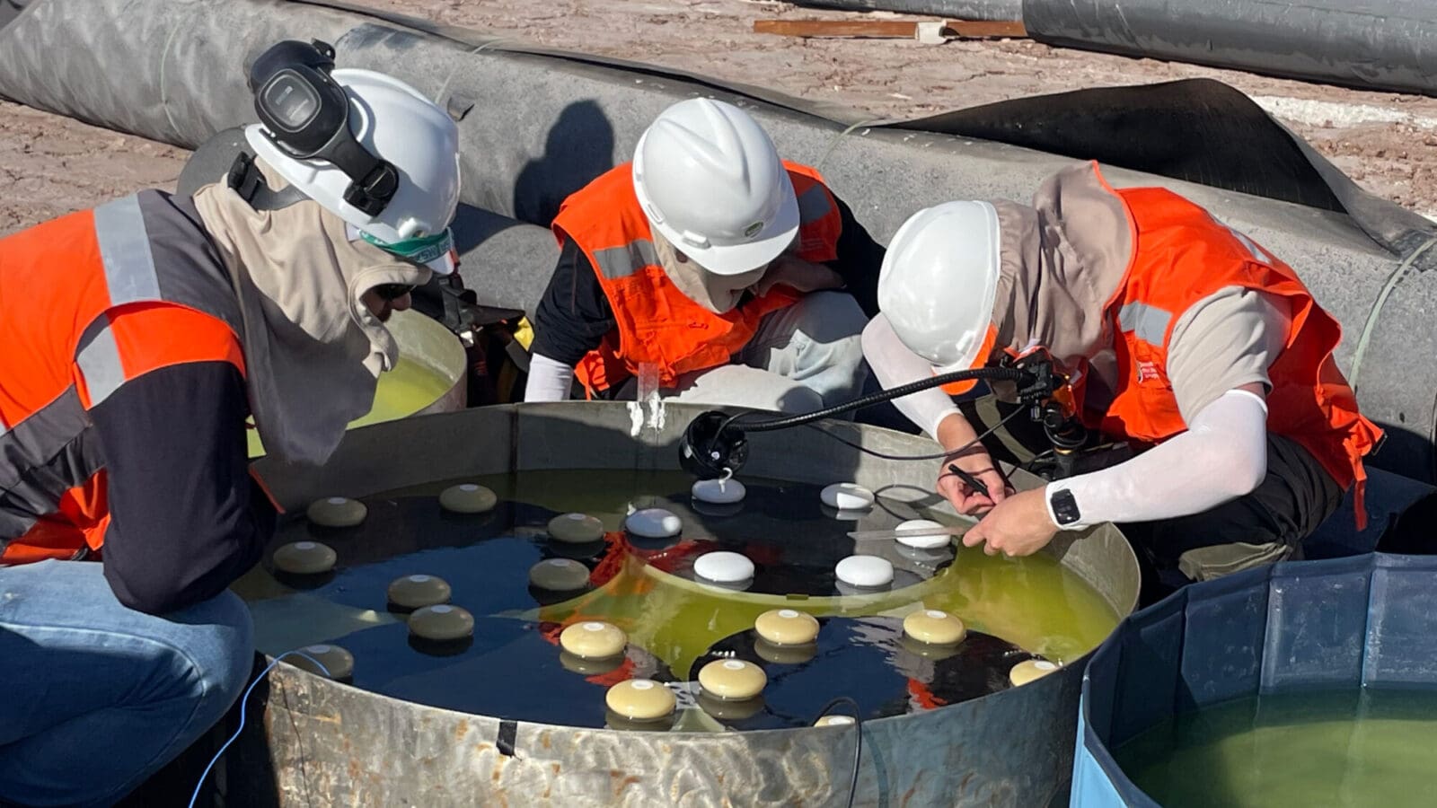 Workers in hard hats and protective gear crouch working around a drum of green liquid