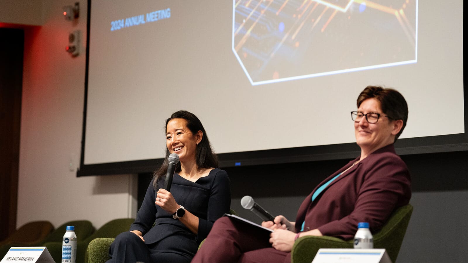 Two women on stage. One speaks into a microphone.