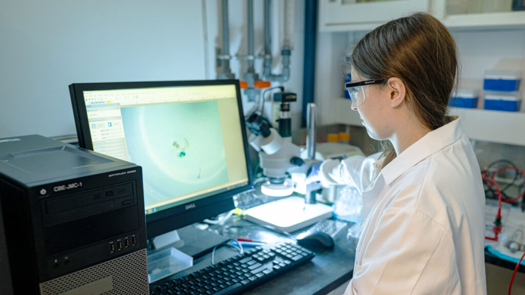 A woman in a lab coat looking at a microscope image on a computer monitor.