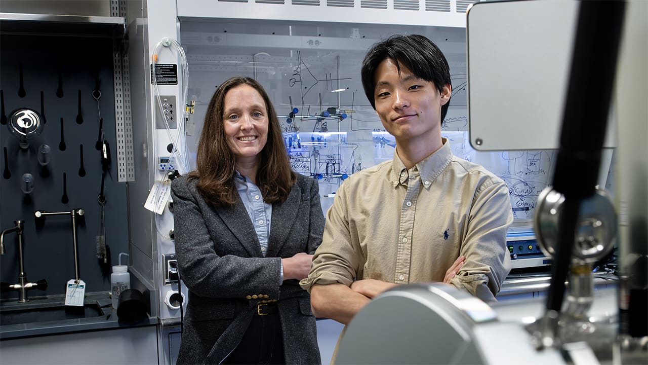 A woman and a man stand side by side in front of a laboratory fume hood.