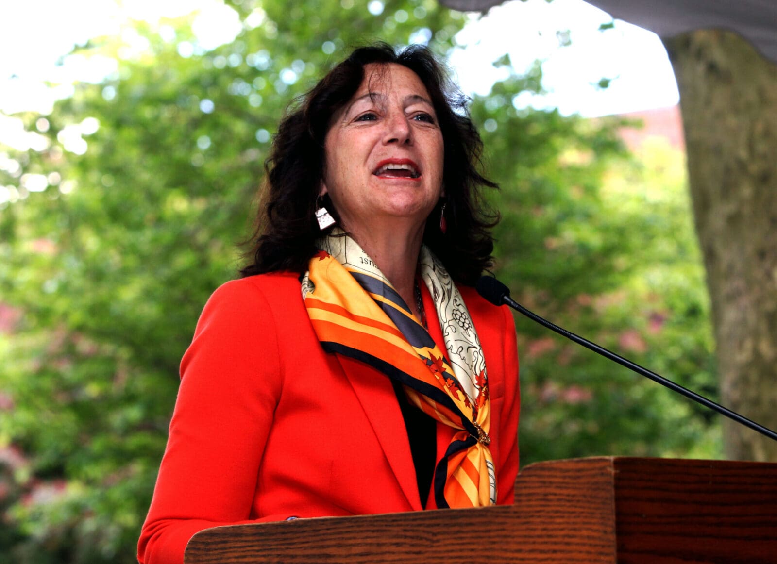 A woman in a reddish orange blazer speaks at a podium outdoors.