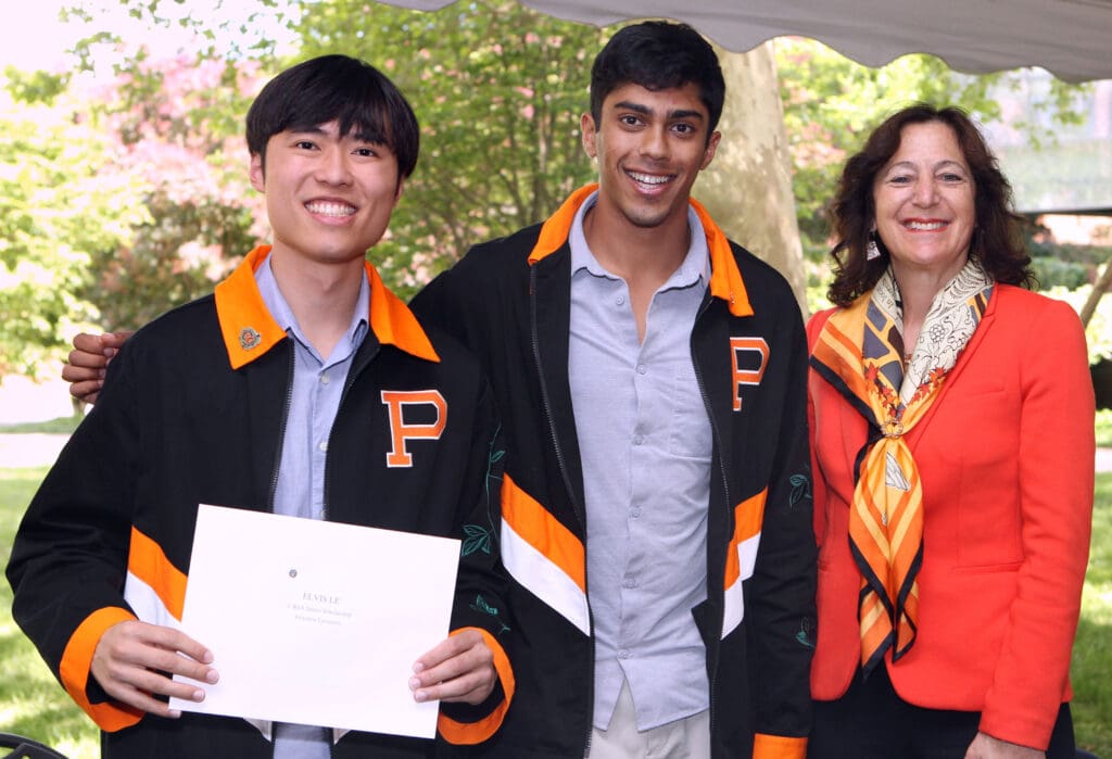 Two students and a professor smiling and posing for a photo under a tent. One student is proudly holding a certificate.