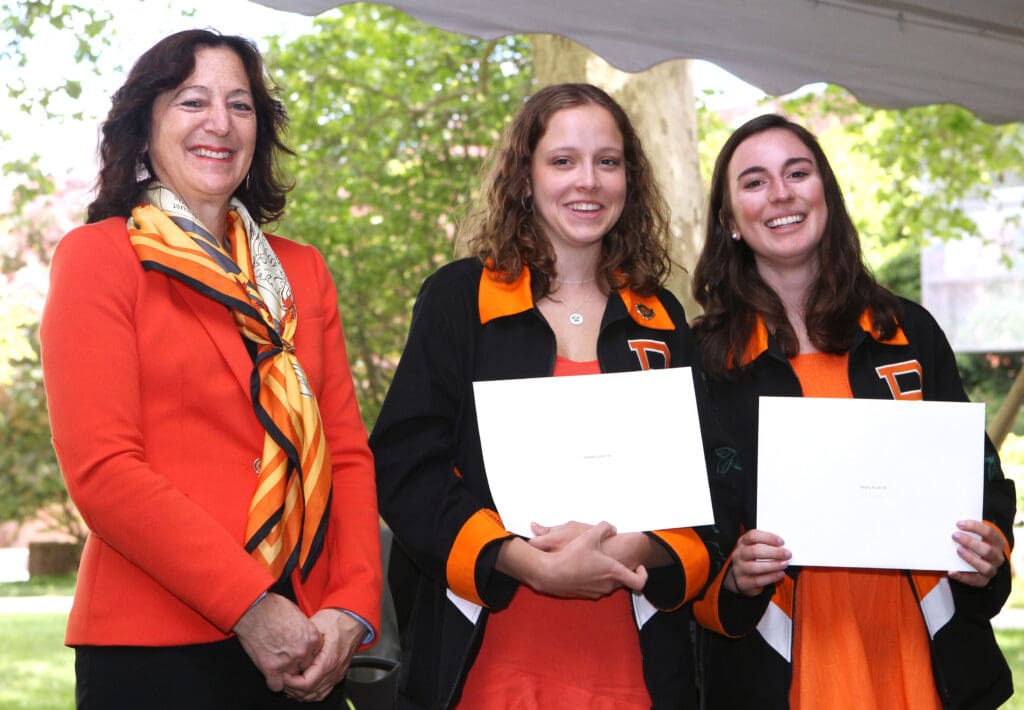 A professor with two students, both holding award certificates, dressed in orange and black outfits, smiling and standing together under a tent outdoors.