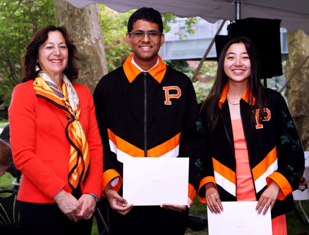 A professor with two students, both holding award certificates, dressed in orange and black outfits, smiling and standing together under a tent outdoors.