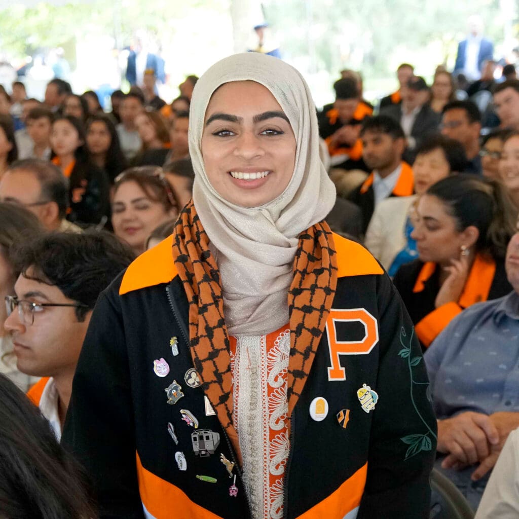 A woman with a bright smile wears an orange and black jacket and beige headscarf while looking at the camera.
