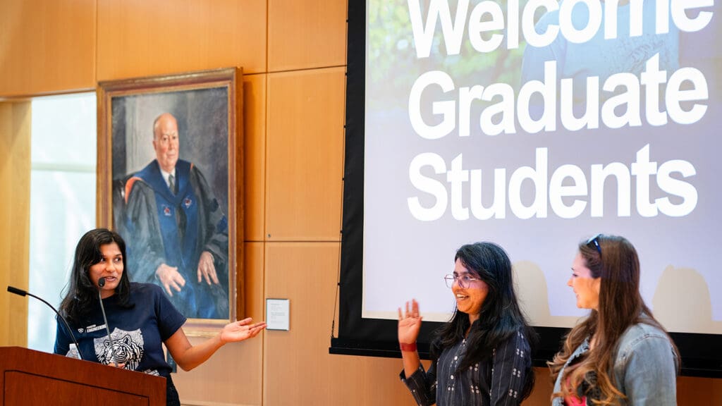 Woman speaking at podium gestures toward two other women standing nearby.