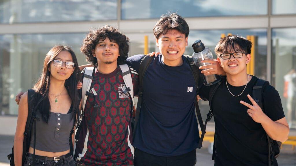 Four students smiling and posing together in front of a glass building facade.