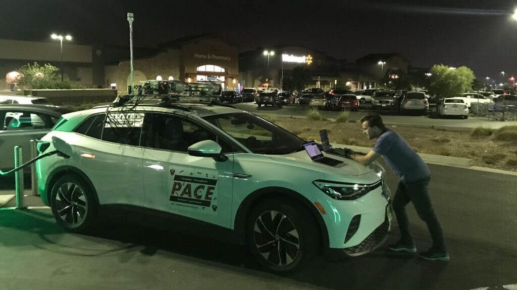 A man works on a laptop open on a car's hood in a parking lot