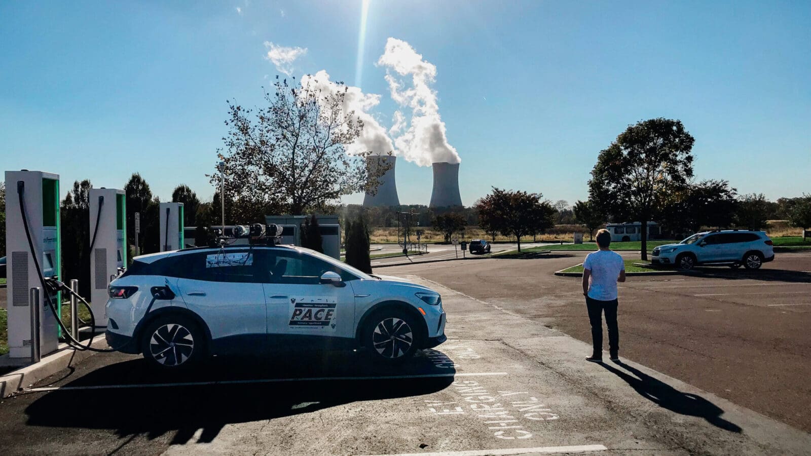 Man stands in front of an electric vehicle at a charging station with cooling towers in the distance