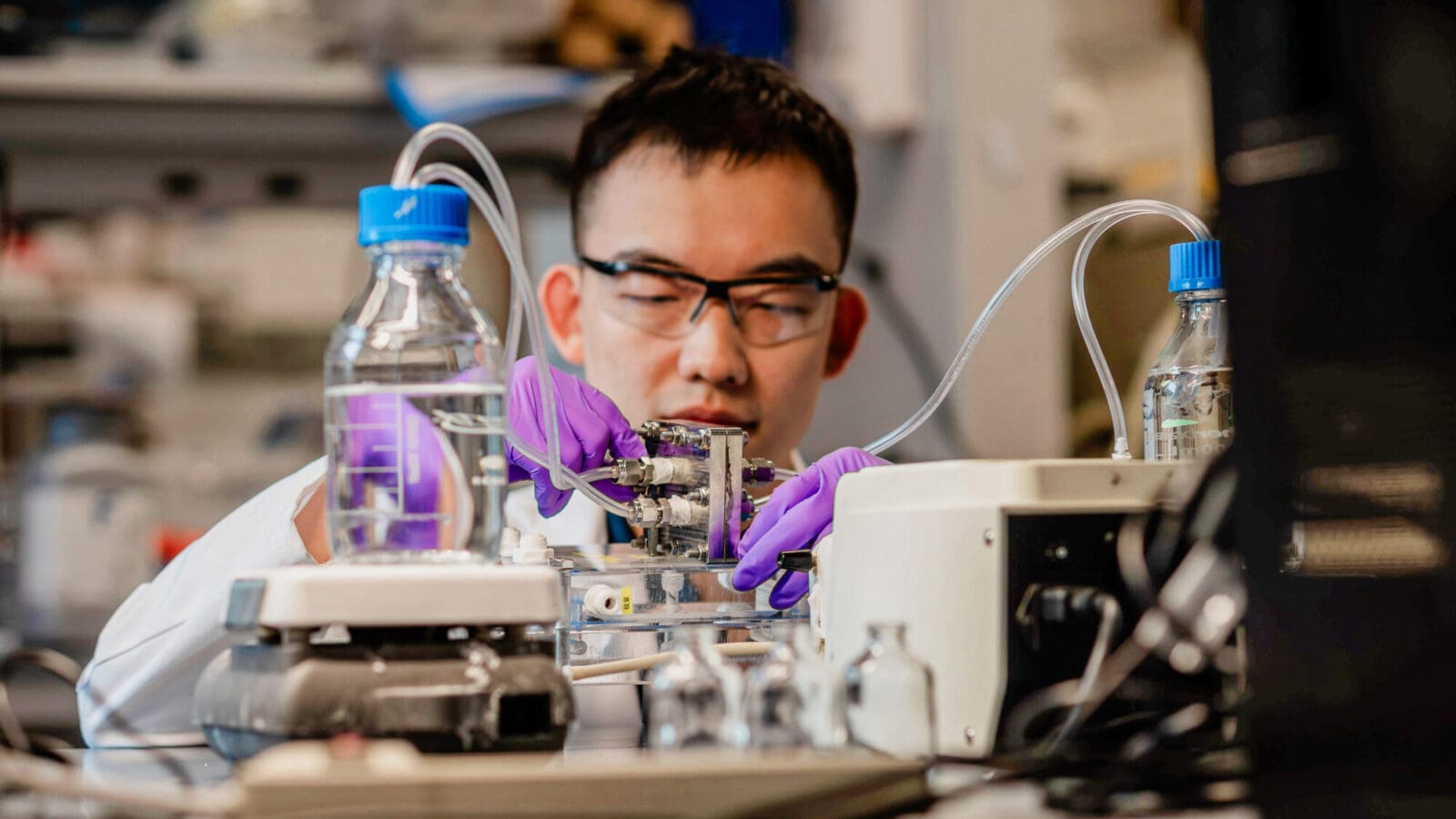A man in a lab coat and safety glasses operates a machine in a laboratory involving bottles of clear liquid and tubing.