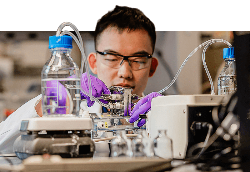 A man in a lab coat and safety glasses operates a machine in a laboratory involving bottles of clear liquid and tubing.