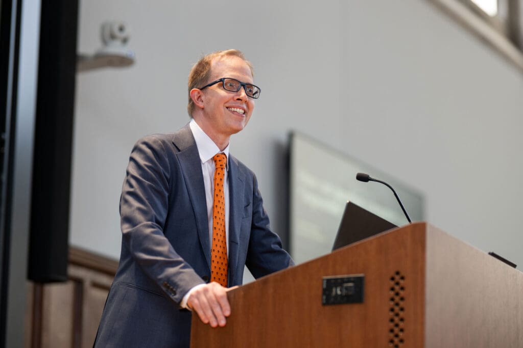 A smiling man in a dark suit and an orange tie stands at a podium.