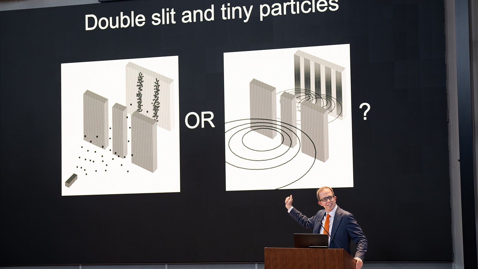 Man at a podium gestures to a screen with a diagram of an experiment.