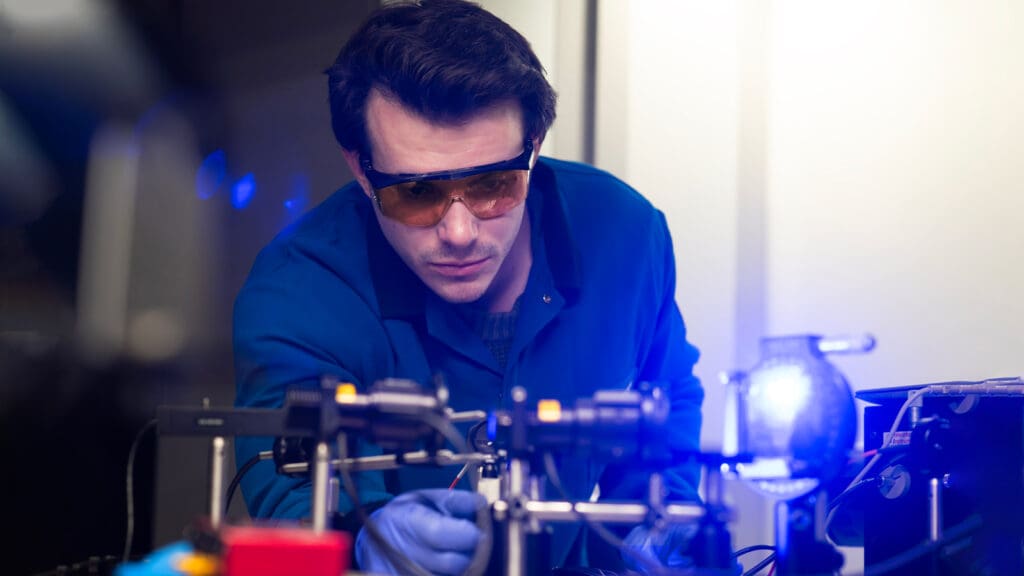 Man in a lab coat leaning over a bench and adjusting optical equipment.