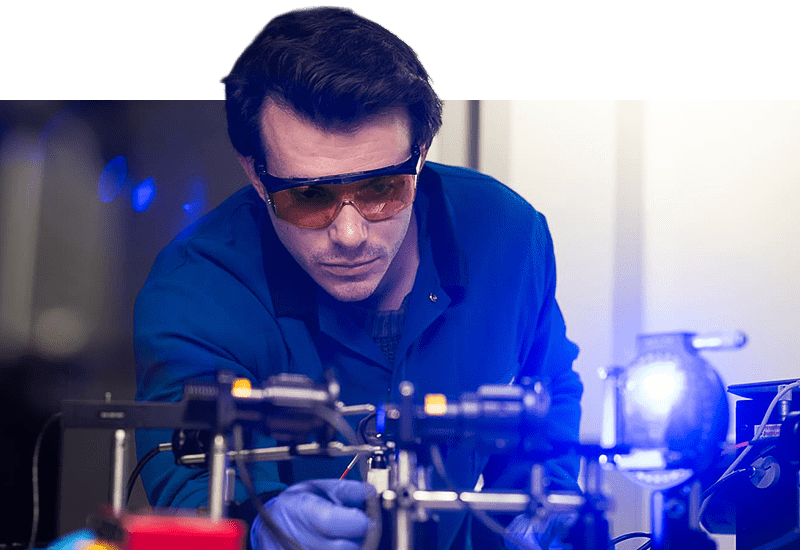 Man in a lab coat leaning over a bench and adjusting optical equipment.